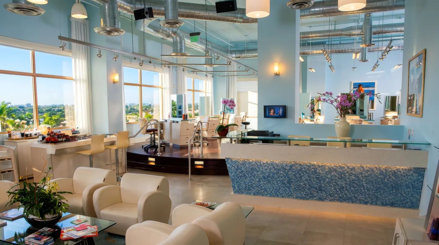 Salon Sora lounge and reception in Boca Raton: white lounge chairs, blue mosaic reception desk, styling stations and tall windows over palm trees (AI-enhanced photo)